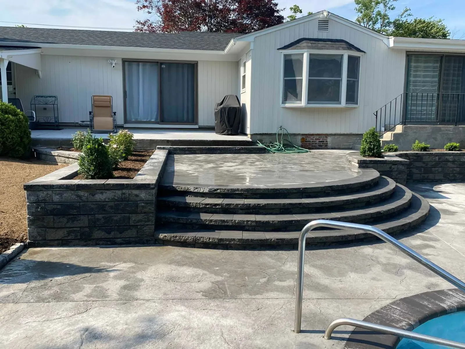 A white house with a sliding glass door and bay window, featuring new tiered grey stone steps leading to a concrete patio.