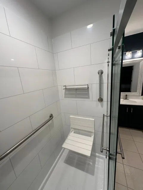 A walk-in shower with white tiled walls, a foldable shower seat, grab bars, a towel rack, and a glass door in a modern bathroom.