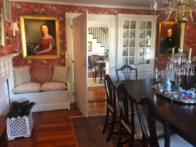 A dining room with red floral wallpaper, portraits, and a table set for a meal.