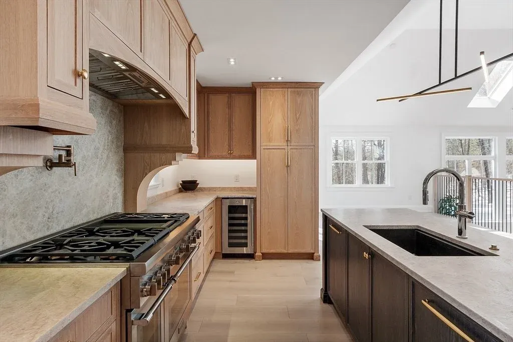 Modern kitchen with light wood cabinets, black kitchen island, marble countertops, stainless steel appliances, and large windows in a bright, open space.