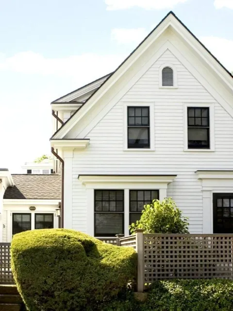 White house with black window frames, a gabled roof, and a small front yard with a trimmed bush and a wooden fence.