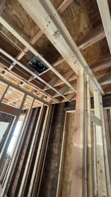 Interior view of a building under construction, showing framing, ceiling beams, and a vent. Wooden structure.