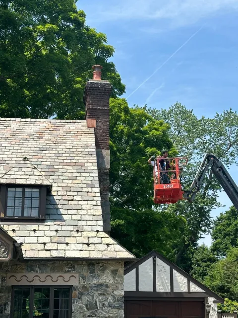 A person in a red aerial lift works on the chimney of a stone house surrounded by trees under a blue sky.
