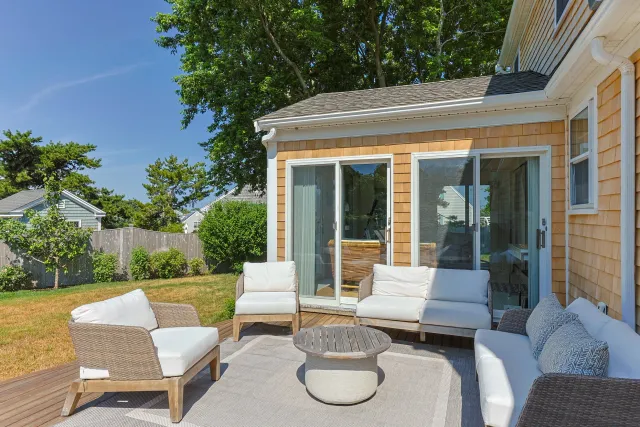 Outdoor patio with beige wicker furniture, white cushions, and a gray rug, adjacent to a house with sliding glass doors and tan wooden siding, under a green leafy tree. Roofing and siding installation by Watkins Works Cape Cod.