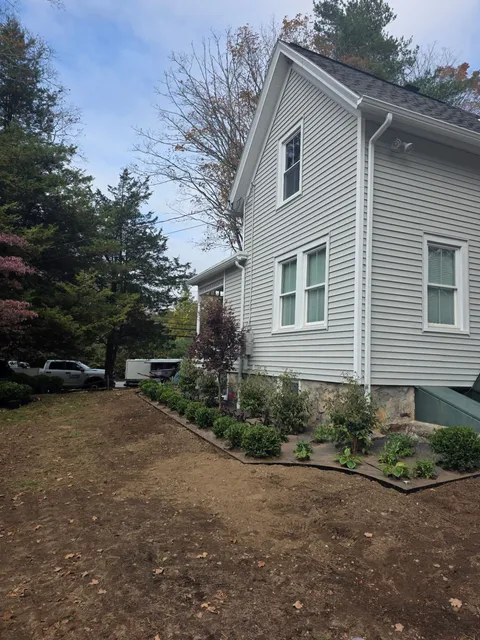 Side view of a light gray house with white trim, set in a yard with a garden bed and trees.