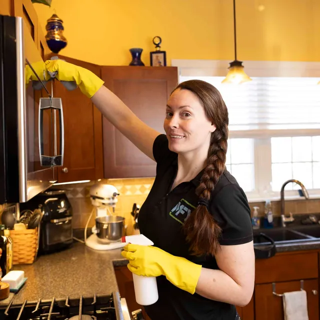 A smiling woman cleaning the top of a microwave while holding a spray bottle