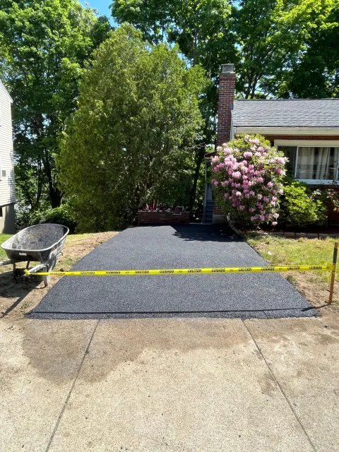 Gravel driveway beside a house with a pink flowering bush and brick chimney, bordered by trees and a concrete path.