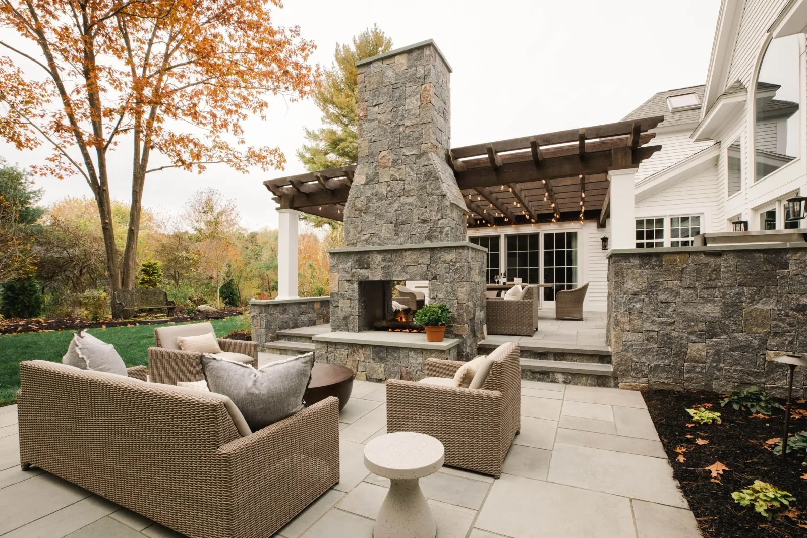 A large patio set across two levels with a feature fireplace in the middle and rattan garden furniture, some of which is covered by a pergola