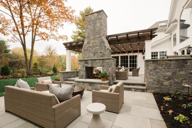 A large patio set across two levels with a feature fireplace in the middle and rattan garden furniture, some of which is covered by a pergola
