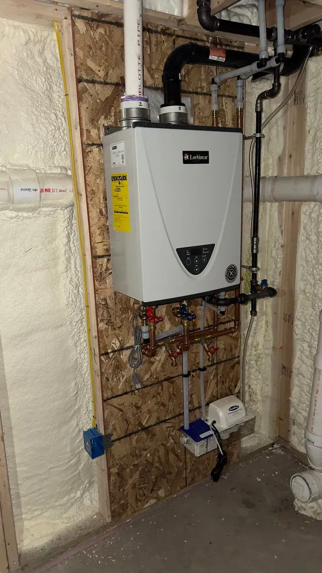 Tankless water heater mounted on a plywood wall in a basement. Pipes and wiring are visible.