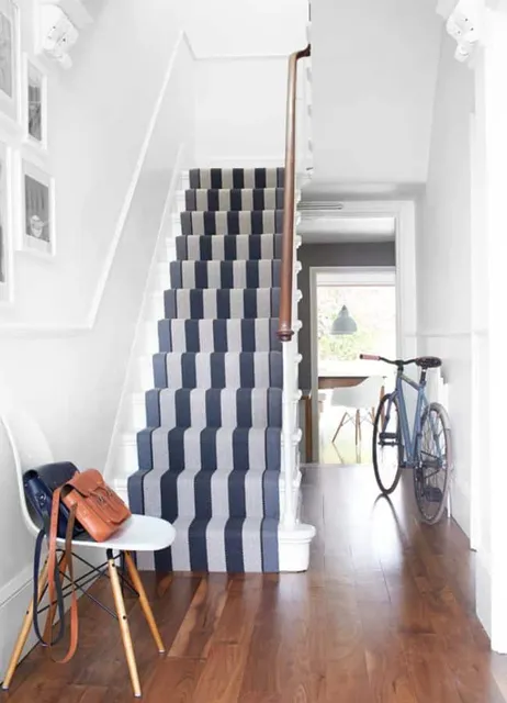 Hallway featuring a stair runner installation with bold blue and grey stripes, a white chair with a tan purse, and a blue bicycle against a white wall. Wood flooring adds warmth to the bright, modern space.