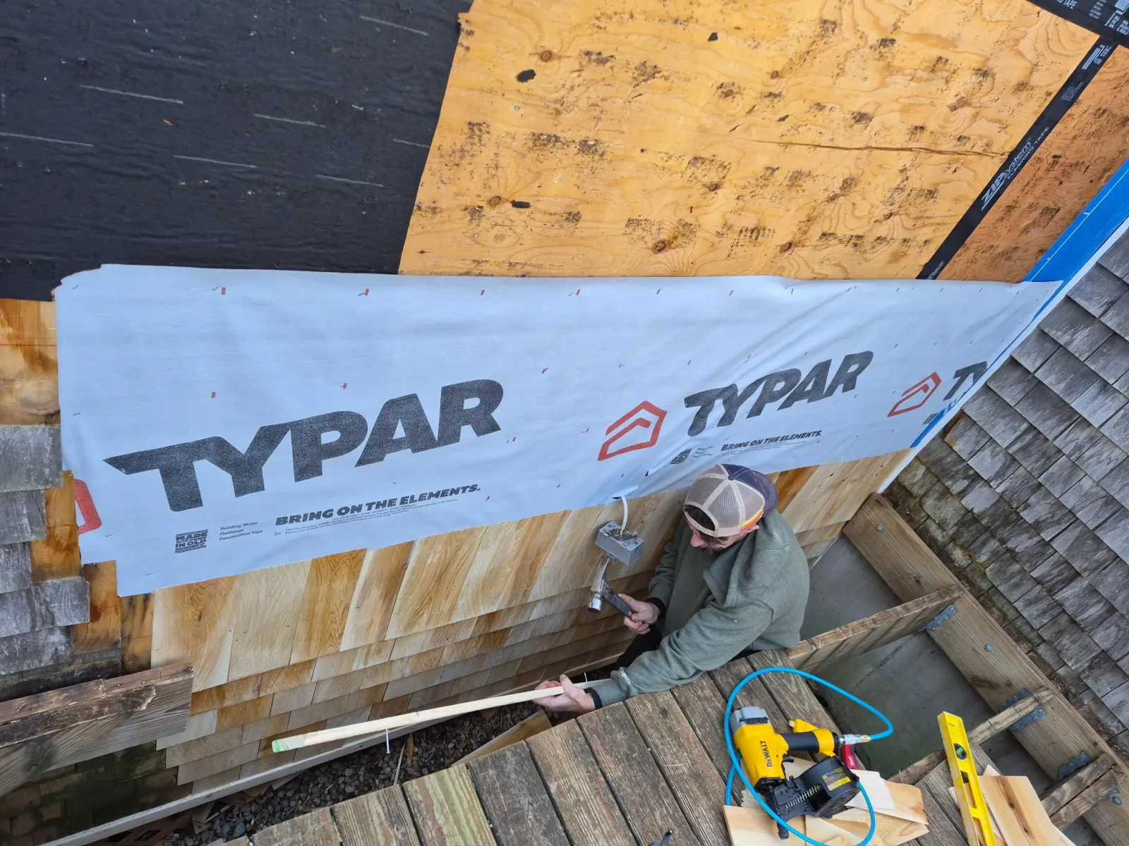 A worker on a scaffolding installs house wrap with the brand name TYPAR, using a hammer and nails, amid wooden siding and construction tools.
