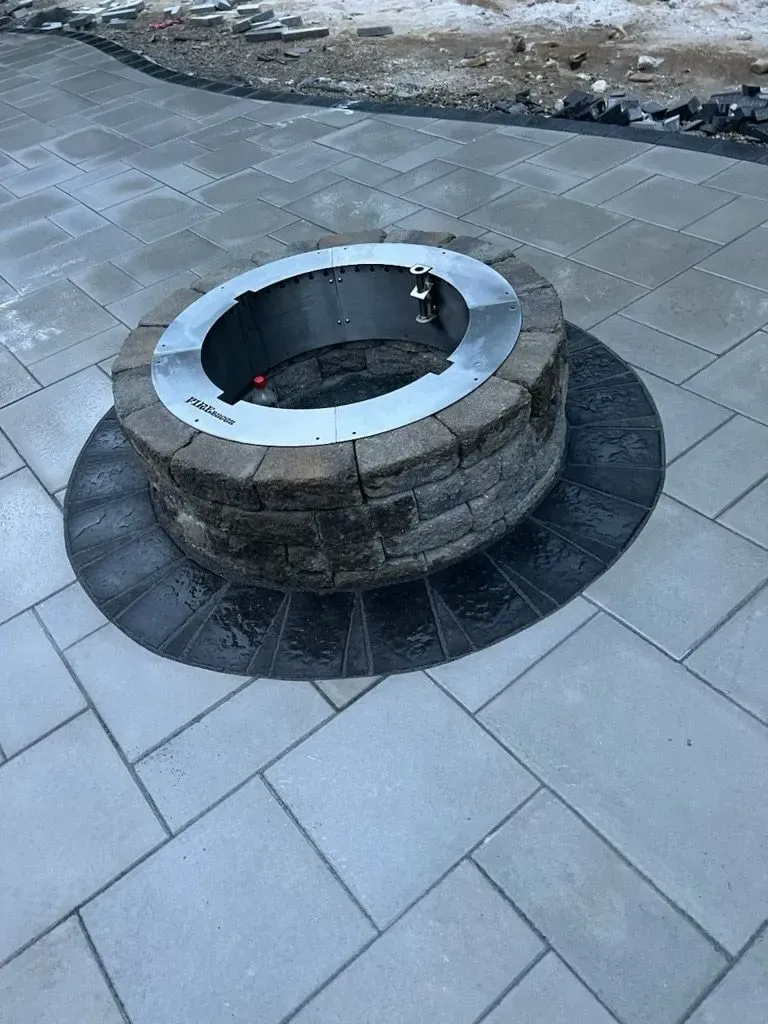 A round stone fire pit with a steel metal insert sits in the center of a paved gray patio.