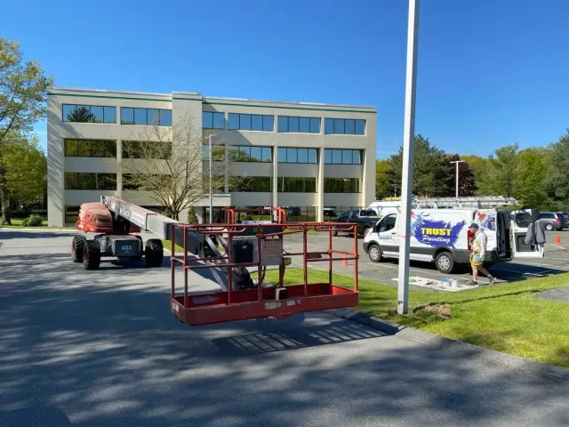 A construction lift sits in a parking lot in front of a three-story office building with a service van parked nearby.