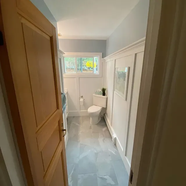 A small, light-walled bathroom with a corner shower stall, a toilet, and patterned floor tile under a window.