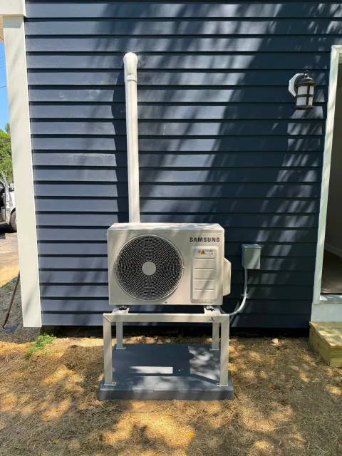 Outdoor HVAC unit against a blue house wall in sunlight, with a vent pipe and utility box.