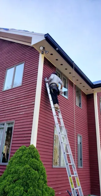 Professional contractor on a ladder performing exterior home repairs on a red farmhouse with vinyl siding.