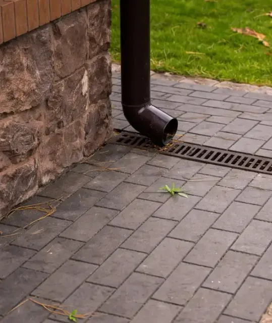 Linear drainage channel installed in a paver walkway next to a stone foundation and downspout