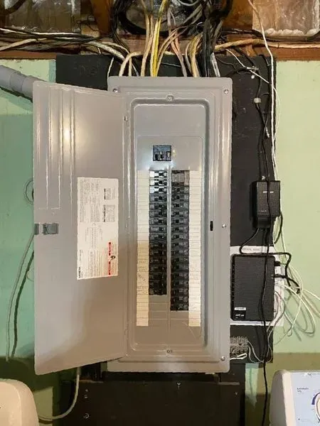 Open electrical panel with numerous circuit breakers, wires overhead, and an attached modem.
