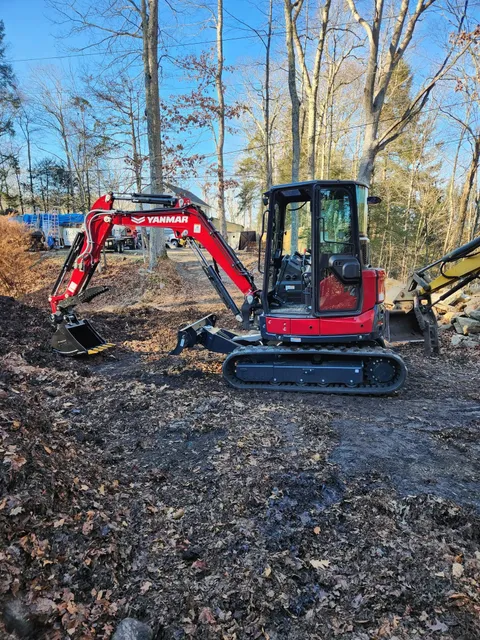 Red excavator parked on a leaf-covered woodland trail under a clear blue sky