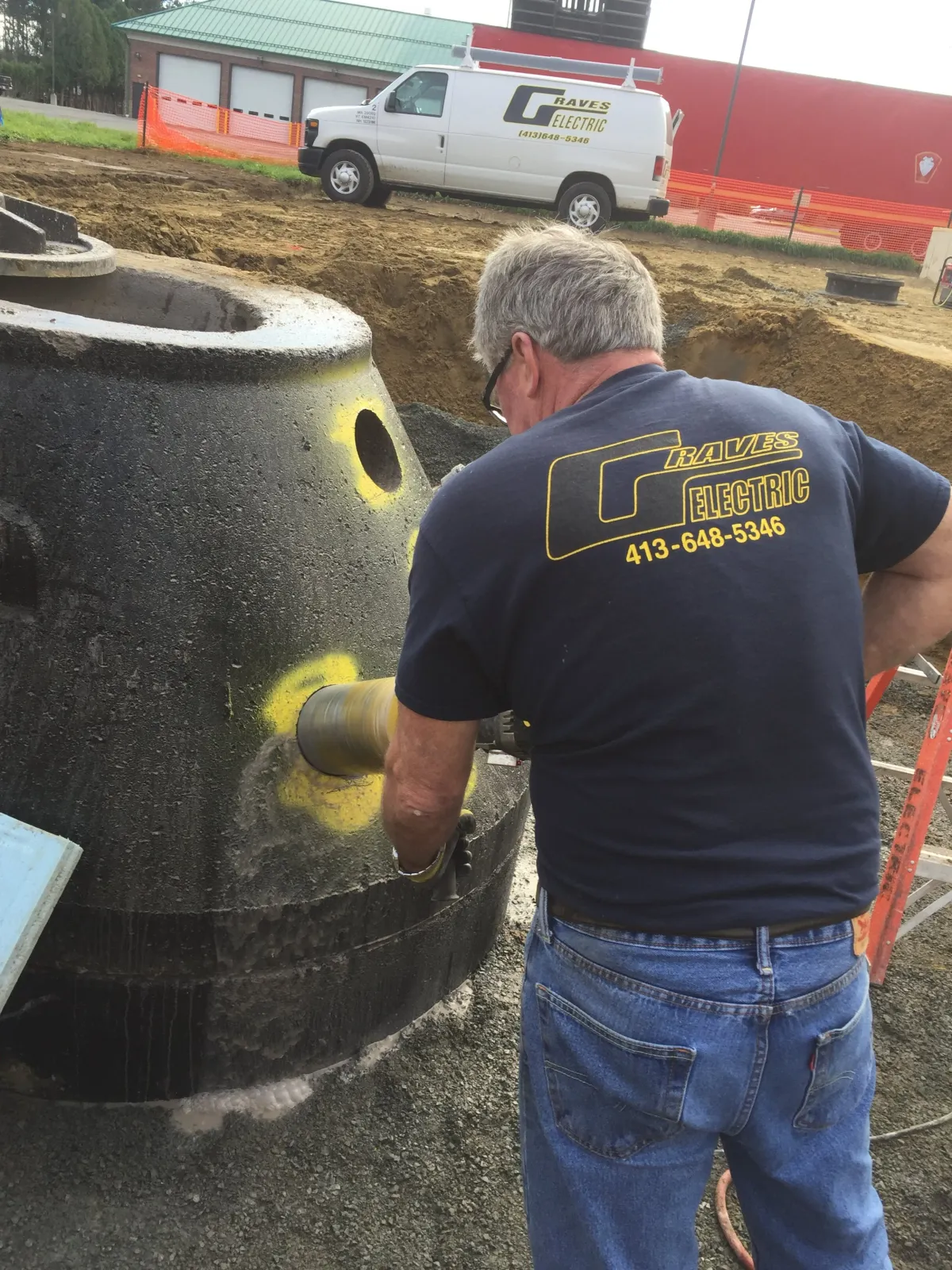 A man in a navy T-shirt and jeans working on a large black underground structure at a construction site. The T-shirt has yellow text and graphics for Raves Electric, and the man is using a power tool on the structure.