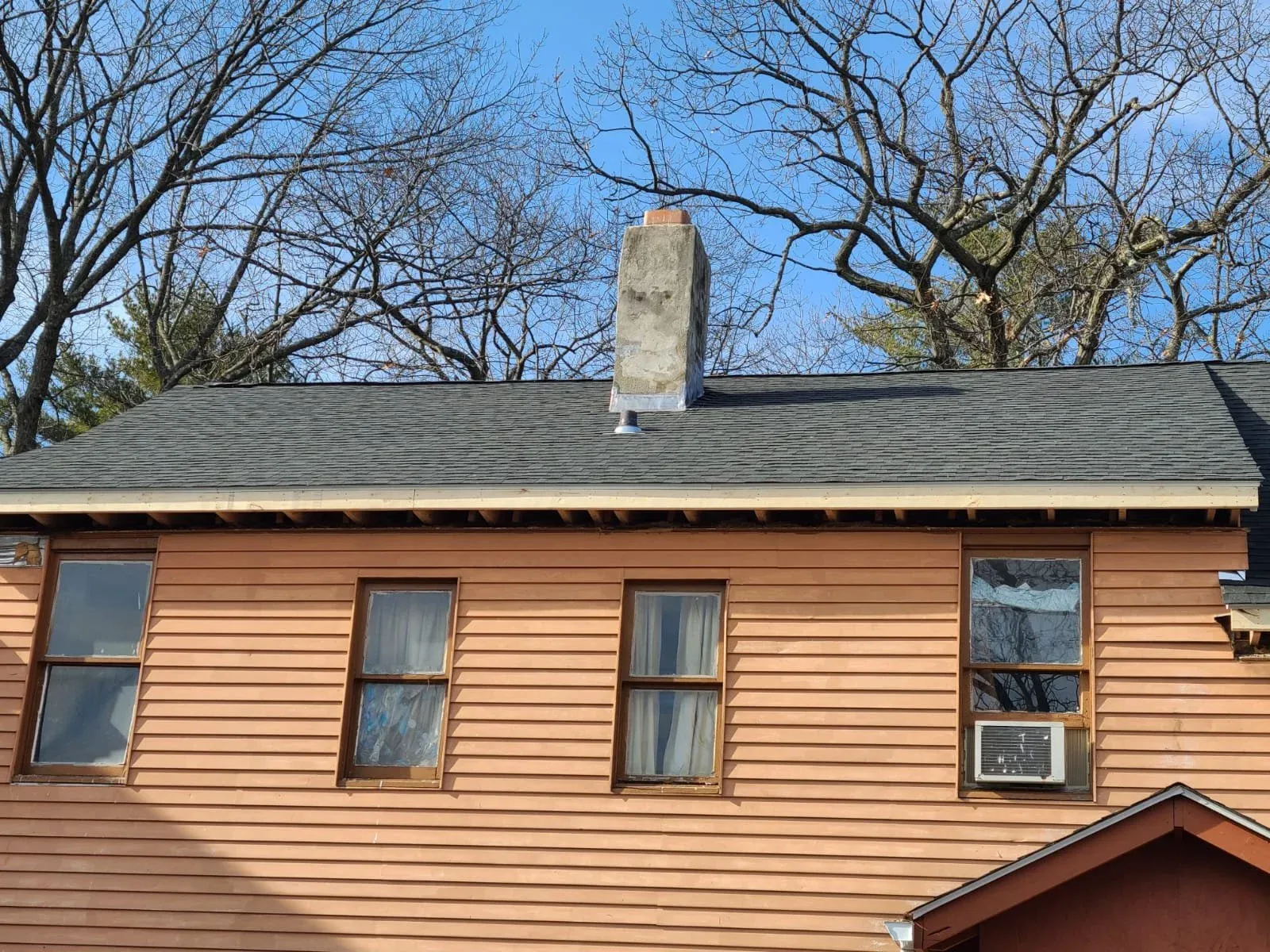 A two-story house with brown siding, four windows on the upper floor, one with an air conditioning unit, a gray shingled roof, and a chimney. Bare trees and a blue sky are in the background.