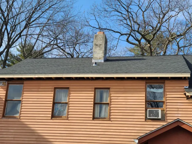 A two-story house with brown siding, four windows on the upper floor, one with an air conditioning unit, a gray shingled roof, and a chimney. Bare trees and a blue sky are in the background.