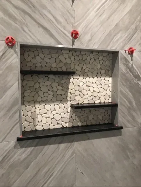 Wall niche filled with small white round stones and two black shelves, mounted on a wall with large gray marble tiles.