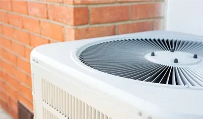 Close-up of an outdoor air conditioning unit next to a brick wall, showing the metal fan grille and the side vents of the unit.