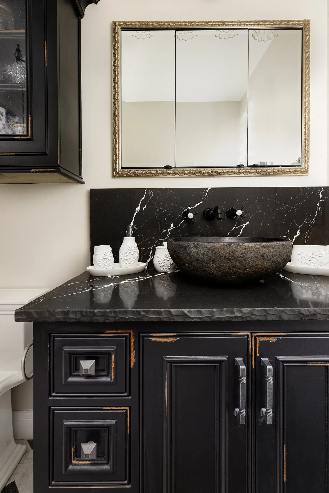 Bathroom vanity area with black distressed cabinetry, black marble countertop, a stone vessel sink, a black backsplash, and a large mirror with ornate gold frame.