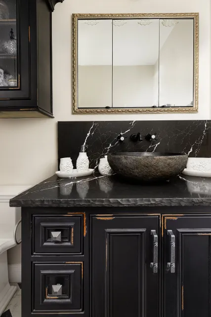 Bathroom vanity area with black distressed cabinetry, black marble countertop, a stone vessel sink, a black backsplash, and a large mirror with ornate gold frame.
