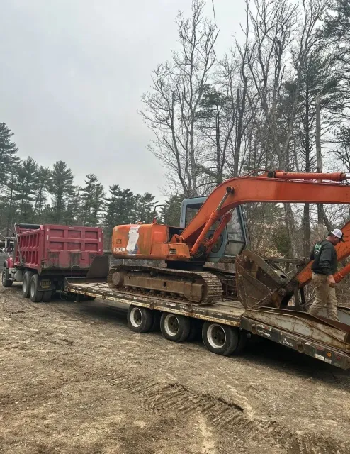 An orange excavator sits on a flatbed trailer attached to a red dump truck at an outdoor construction site.