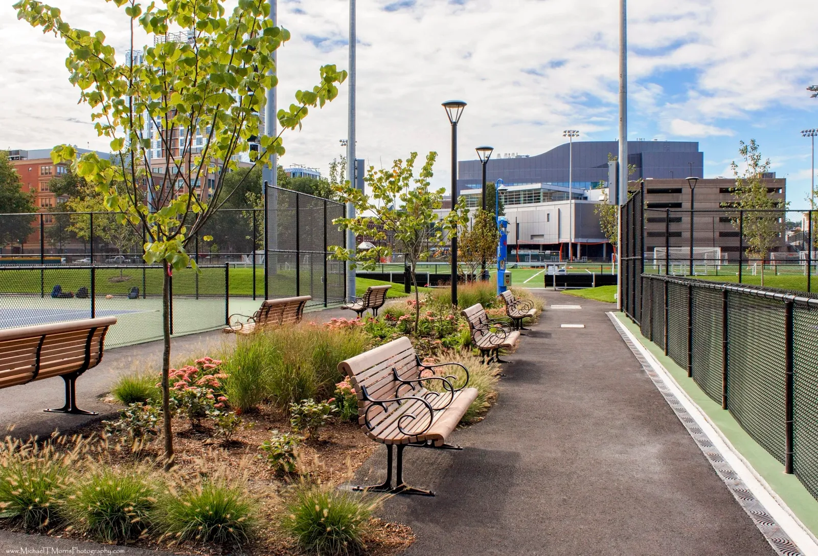 Beautifully landscaped urban park walkway in Massachusetts with stylish benches, manicured greenery, and tennis courts — a showcase of Xquisite Landscaping’s expert commercial landscape design.