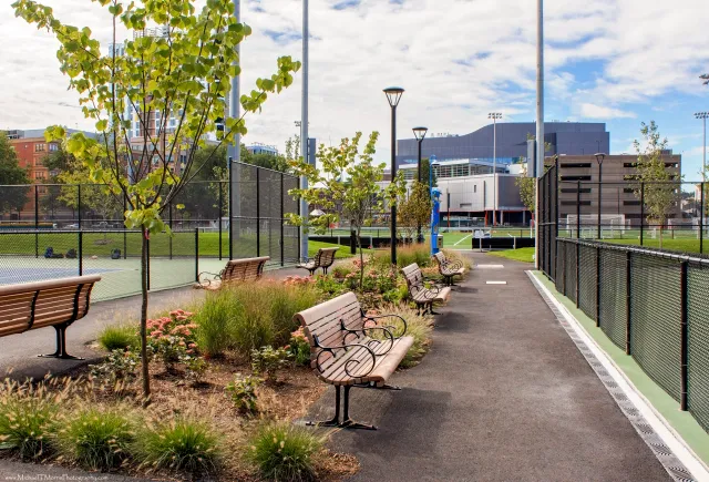 Beautifully landscaped urban park walkway in Massachusetts with stylish benches, manicured greenery, and tennis courts — a showcase of Xquisite Landscaping’s expert commercial landscape design.