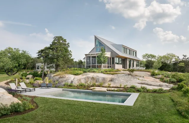 Modern house by a maine architect sits on a rocky hill surrounded by greenery, with a rectangular pool and lounge chairs on the patio. Design-build elements shine under partly cloudy skies.