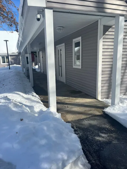 Covered walkway with snow on either side of the path, leading to a double door. Gray siding, white pillars.