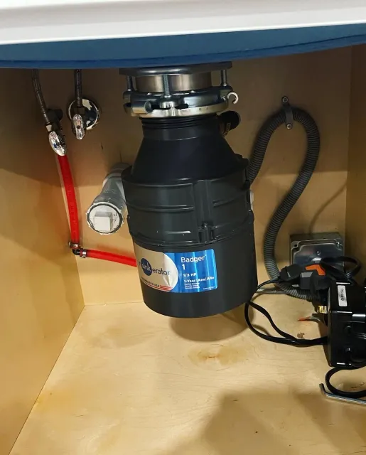 Under-sink plumbing with a garbage disposal unit, red water supply pipe, flexible electrical cord, and drainage hoses in a kitchen cabinet.