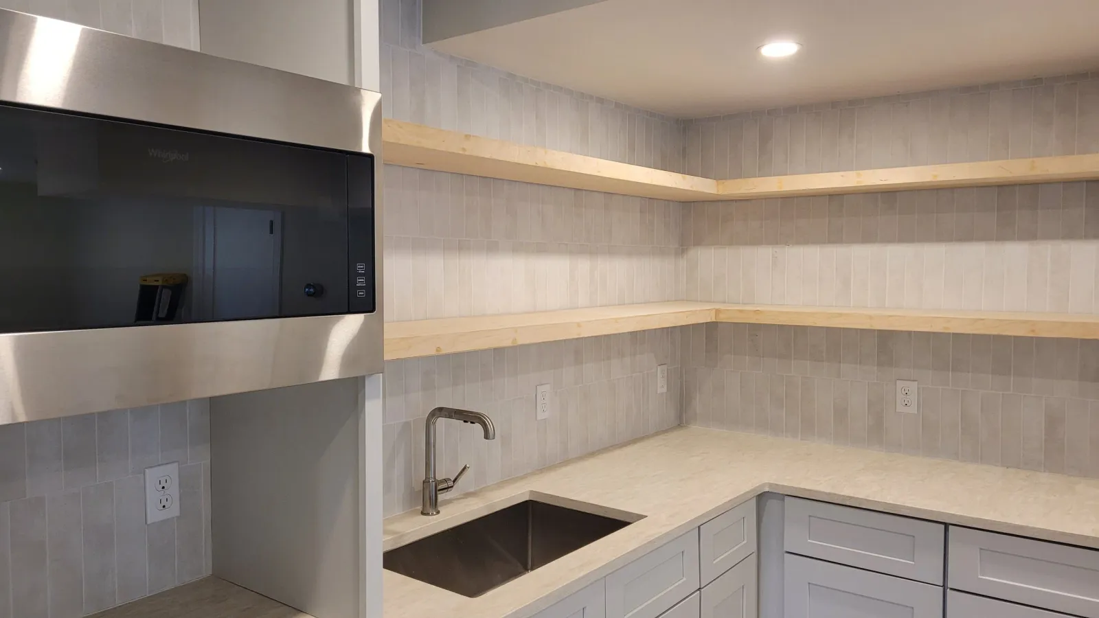 Empty kitchen with light-colored cabinets, a small sink, beige countertop, two open wooden shelves, and multiple electrical outlets with gray tile backsplash.