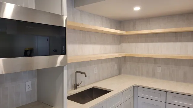 Empty kitchen with light-colored cabinets, a small sink, beige countertop, two open wooden shelves, and multiple electrical outlets with gray tile backsplash.