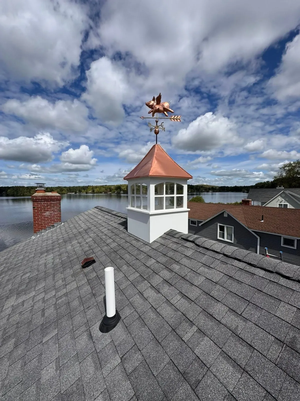 Rooftop weather vane on a white cupola above gray shingles, with a cloudy sky and water in the background