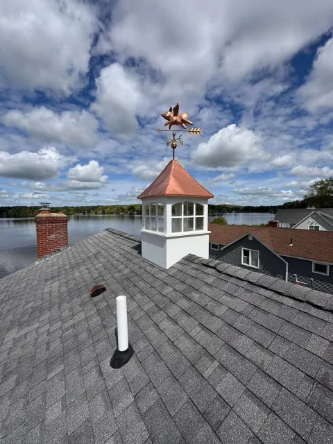 Rooftop weather vane on a white cupola above gray shingles, with a cloudy sky and water in the background
