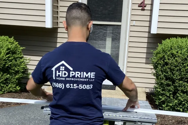 Company owner in a dark blue shirt with 'HD Prime Home Improvement LLC' logo and phone number, working with a saw outside to cut trim.