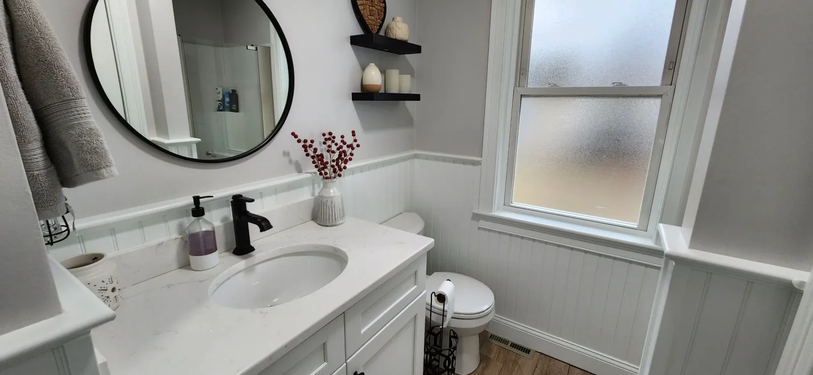 Bathroom with white cabinets, round mirror, and black fixtures. A vase with red berries is on the counter, and floating shelves hold decorative items. A frosted window and toilet are visible.