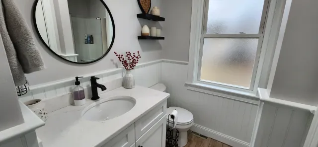 Bathroom with white cabinets, round mirror, and black fixtures. A vase with red berries is on the counter, and floating shelves hold decorative items. A frosted window and toilet are visible.