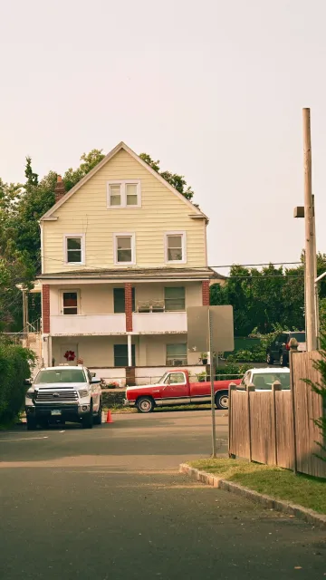 A three-story yellow house with a porch, cars parked in front, and a wooden fence on the side.