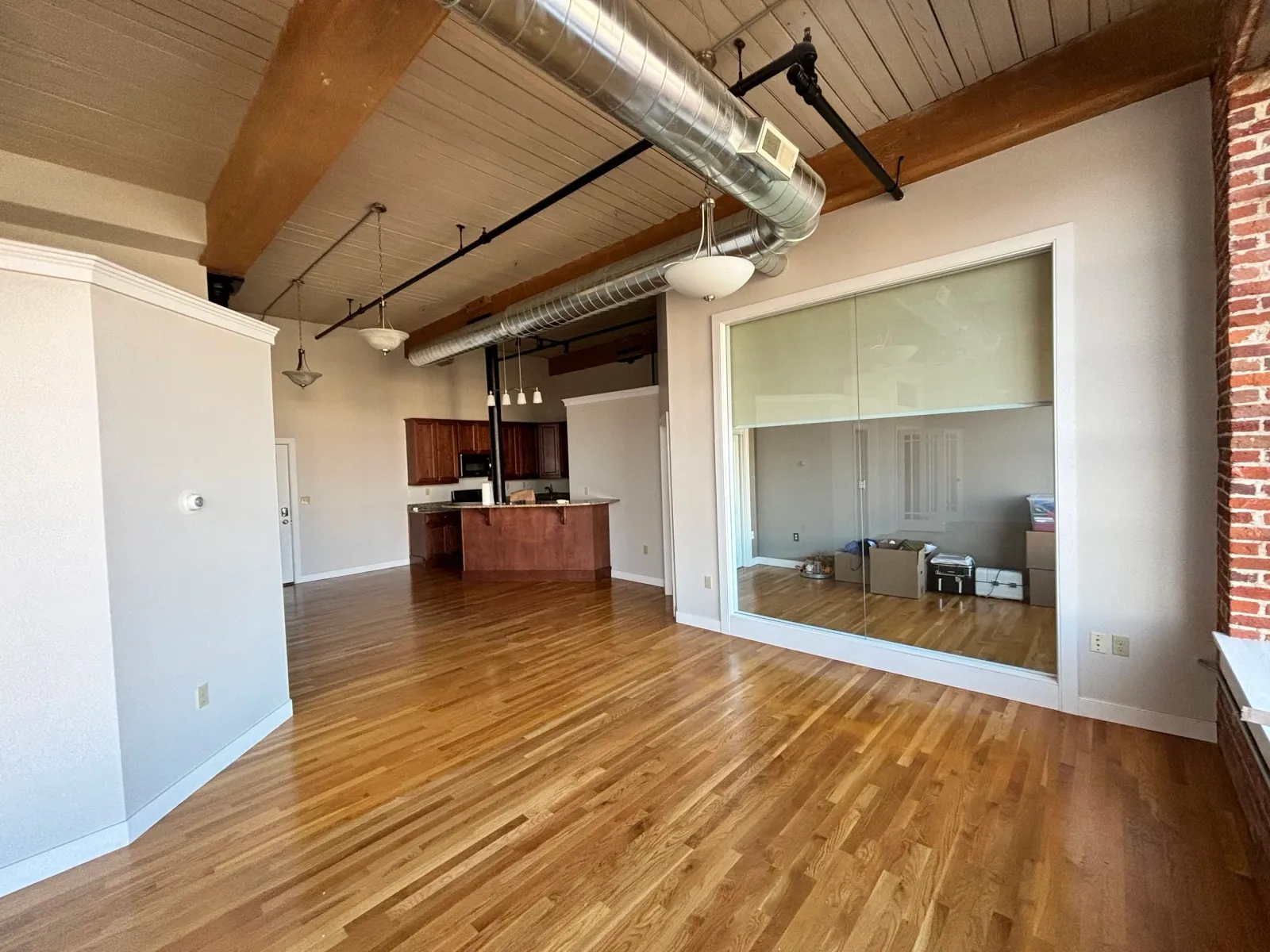 Spacious loft interior with wooden floor, exposed ductwork, kitchen area with wooden cabinets, and a large window.