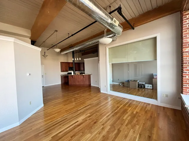 Spacious loft interior with wooden floor, exposed ductwork, kitchen area with wooden cabinets, and a large window.