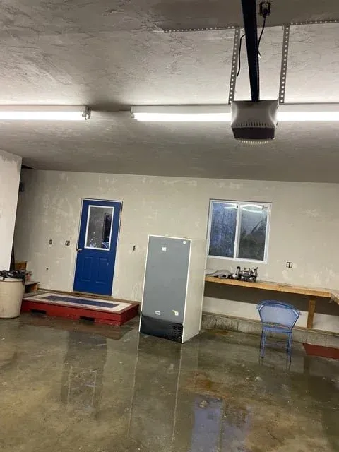 Interior of a garage or workshop with a blue door, window, workbench, and a standing appliance or cabinet, with fluorescent lighting and a concrete floor.