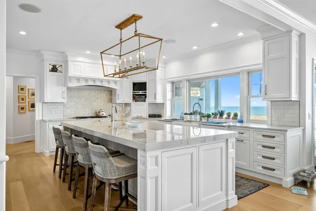 Custom kitchen remodel at William Crowell Road in Dennis, MA, featuring a large white marble island, woven bar stools, gold pendant lights, white cabinetry, and ocean views through large windows