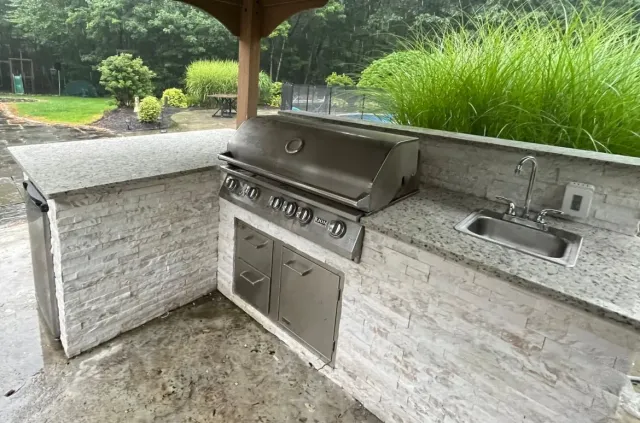 Outdoor built-in stone grill and sink under a covered patio, with greenery in the background